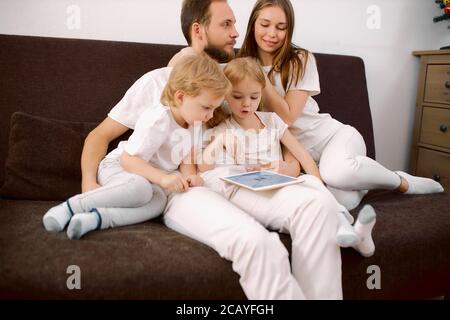 la jeune famille caucasienne était composée d'enfants, de petite fille, de fils et de parents assis sur un canapé portant des vêtements décontractés, en utilisant une tablette de gadget moderne Banque D'Images