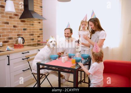 drôle de famille caucasienne passer du temps, célébrer l'anniversaire avec le chien à la maison. Parents, enfants et animaux domestiques - animaux de compagnie en casquettes d'anniversaire, décontractés Banque D'Images