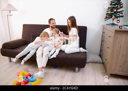 la famille heureuse se composait de mère, père, fille et fils, assis sur un canapé portant des vêtements blancs, utilisant une tablette, des technologies modernes Banque D'Images