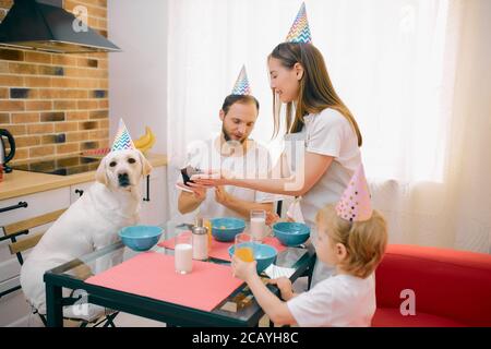 drôle de famille caucasienne passer du temps, célébrer l'anniversaire avec le chien à la maison. Parents, enfants et animaux domestiques - animaux de compagnie en casquettes d'anniversaire, décontractés Banque D'Images