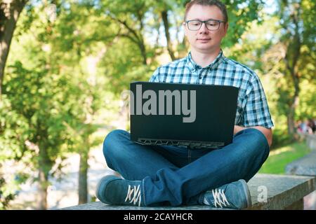 Le gars est assis dans le parc et travaille à un ordinateur portable. Banque D'Images