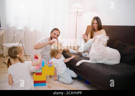 famille caucasienne amicale ont un animal de compagnie ludique, chien blanc à la maison. homme, femme et enfants dans la salle de séjour, jouer avec les enfants et avec le chien, portant décontracté Banque D'Images