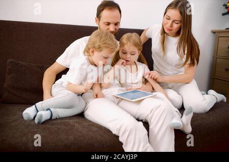 la famille heureuse se composait de mère, père, fille et fils, assis sur un canapé portant des vêtements blancs, utilisant une tablette, des technologies modernes Banque D'Images