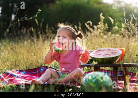 une petite fille drôle sur un pique-nique d'été mange un rouge mûr délicieux melon d'eau juteux Banque D'Images