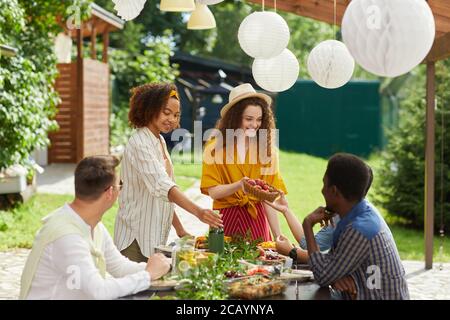 Groupe multiethnique de personnes appréciant dîner sur la terrasse extérieure en été, se concentrer sur la jeune femme souriante donnant des fruits frais et des baies à travers la table, Banque D'Images