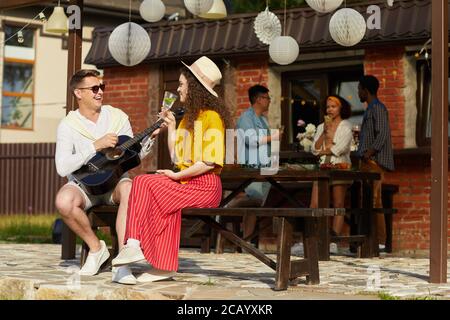 Portrait grand angle des jeunes qui profitent de la fête en plein air en été avec l'accent sur l'homme jouant de la guitare à l'amie en premier plan, espace de copie Banque D'Images