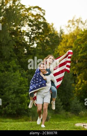 Portrait vertical en longueur de jeune couple insouciant qui court vers un appareil photo sur une pelouse verte avec drapeau américain agité Banque D'Images