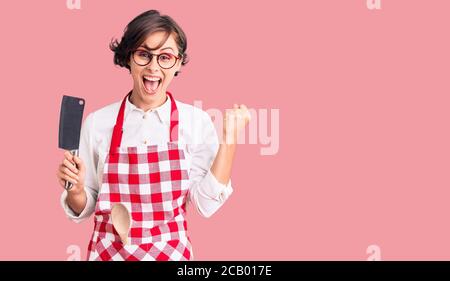 Belle jeune femme aux cheveux courts portant un tablier boulanger professionnel tenant le couteau criant fier, célébrant la victoire et le succès très excitée d'esprit Banque D'Images