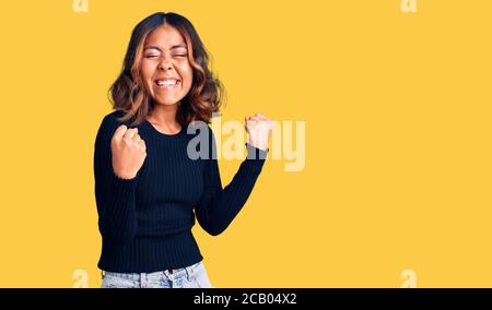 Jeune femme de course mixte, portant des vêtements décontractés très heureux et excités faisant le geste gagnant avec les bras levés, souriant et criant pour des suces Banque D'Images