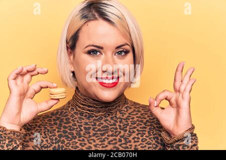 Jeune belle blonde plus grande femme de taille mangeant français dessert macaron sur fond jaune faire signe ok avec les doigts, souriant sympathique gestuing exc Banque D'Images