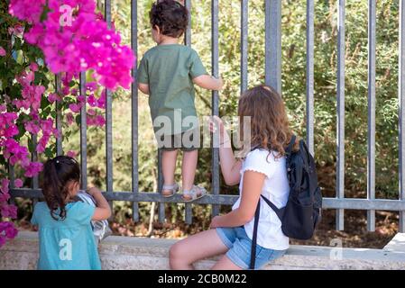 Les petites filles aux cheveux blonds et aux cheveux noirs et leur petit frère aux cheveux bouclés regardent à travers une clôture métallique fermée. Divers enfants vont à l'école. Banque D'Images