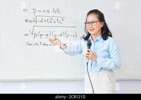 Femme asiatique mature enseignant de mathématiques parlant dans le microphone lors de la demande l'étudiant doit résoudre l'équation sur le tableau blanc Banque D'Images