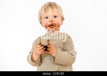 Adorable tout-petit blond mangeant une barre de chocolat avec grand plaisir, fond blanc, Pologne Banque D'Images