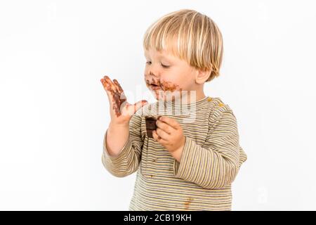 Adorable tout-petit blond mangeant une barre de chocolat avec grand plaisir, fond blanc, Pologne Banque D'Images