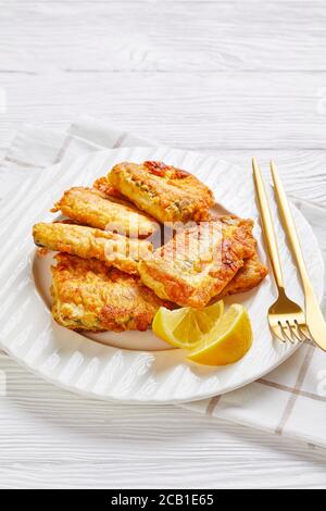 Filet de poisson grillé et croustillant, servi sur une assiette blanche avec des couverts dorés aux quartiers de citron sur un fond de bois blanc avec une serviette, gros plan Banque D'Images