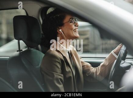 Femme d'affaires avec des écouteurs conduisant sa voiture au bureau et souriant. Femme qui écoute de la musique tout en conduisant au travail. Banque D'Images