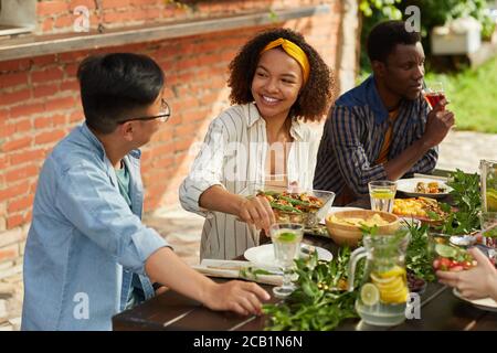 Portrait d'une femme afro-américaine souriante tenant un plat de pommes de terre tout en appréciant le dîner avec des amis et la famille à l'extérieur à la fête d'été, espace de copie Banque D'Images