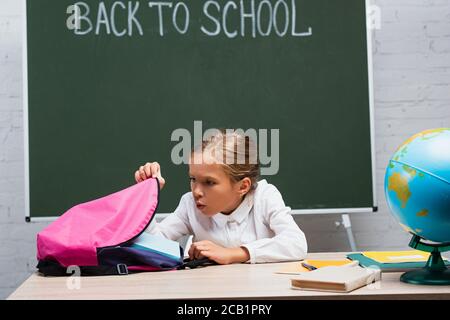 surprise écolière regardant dans un sac à dos tout en étant assis près du bureau globe et tableau noir avec lettrage de retour à l'école Banque D'Images