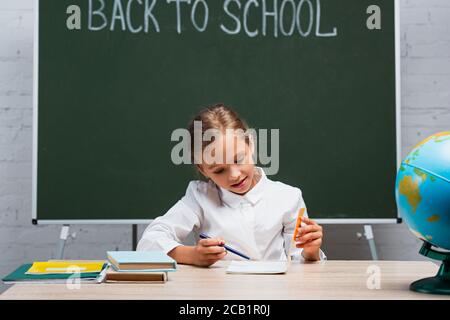 écolière attentive regardant dans un photolivre tout en étant assise au bureau près des manuels scolaires et tableau noir avec inscription rentrée scolaire Banque D'Images