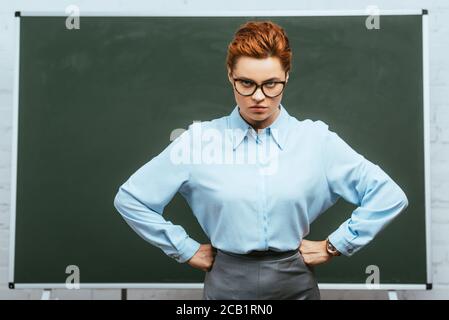 professeur strict dans les lunettes regardant la caméra tout en se tenant avec mains sur les hanches près du tableau noir Banque D'Images