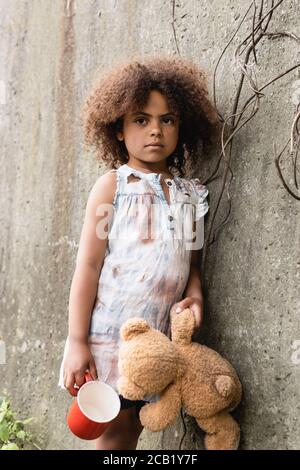 Mendiant enfant afro-américain tenant l'ours en peluche et la tasse près mur en béton sur la rue urbaine Banque D'Images