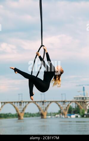 Une femme aerialiste interprète des éléments acrobatiques en accrochant le cerceau aérien sur fond de rivière, de pont et de ciel bleu. Banque D'Images