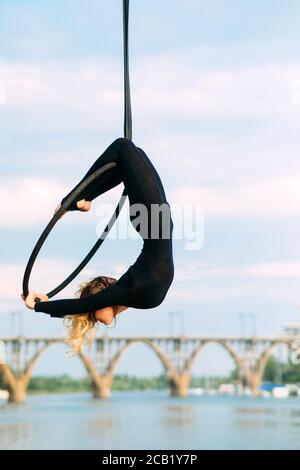 Une femme aerialiste interprète des éléments acrobatiques en accrochant le cerceau aérien sur fond de rivière, de pont et de ciel bleu. Banque D'Images