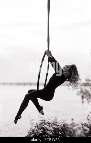 Une femme aerialiste interprète des éléments acrobatiques en accrochant le cerceau aérien contre le fond de la rivière. Image en noir et blanc. Banque D'Images