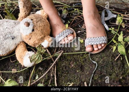 Vue rognée de l'ours en peluche sale près des pieds du pauvre enfant afro-américain au sol en plein air Banque D'Images