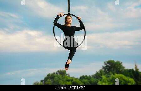 Une femme aerialiste interprète des éléments acrobatiques dans un panier suspendu sur fond de ciel bleu, de nuages blancs et d'arbres. Banque D'Images