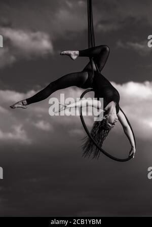 Une femme aerialiste interprète des éléments acrobatiques en accrochant le cerceau aérien sur fond de ciel et de nuages. Image en noir et blanc. Banque D'Images