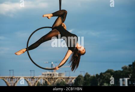 Une femme aerialiste interprète des éléments acrobatiques en accrochant le cerceau aérien sur fond de pont, de ciel bleu et d'arbres. Banque D'Images