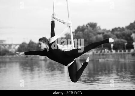 Femme aerialiste effectue des tours acrobatiques sur la suspension de soie aérienne sur fond de rivière, de ciel et d'arbres. Image en noir et blanc. Banque D'Images