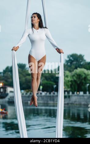 Femme aerialiste effectue des tours acrobatiques sur la suspension de soie aérienne sur fond de rivière, de ciel et d'arbres. Banque D'Images