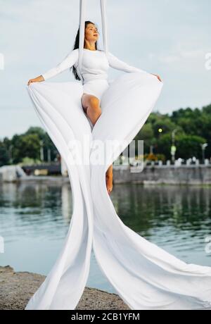 Femme aerialiste effectue des tours acrobatiques sur la suspension de soie aérienne et la pose de tissus sur fond de rivière, de ciel et d'arbres. Banque D'Images