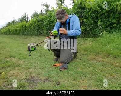 Homme senior avec coupe-brosse sur la pelouse contre le arrière-plan des bagues dans les vêtements de travail et les lunettes de protection Banque D'Images