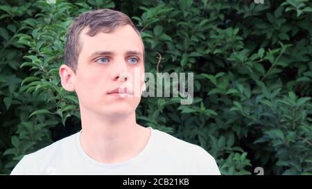 Portrait d'un jeune homme très beau avec des yeux bleus et les cheveux blonds regardant sur le côté contre un arrière-plan de verdure Banque D'Images