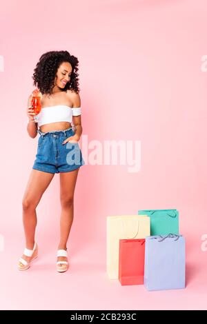 bonne femme afro-américaine tenant un cocktail et debout à la main dans la poche tout en regardant les sacs de shopping sur rose Banque D'Images