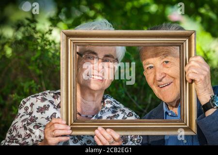 Vue frontale d'un couple en train de rire en retraite regardant à travers un cadre d'image devant une haie floue. Banque D'Images