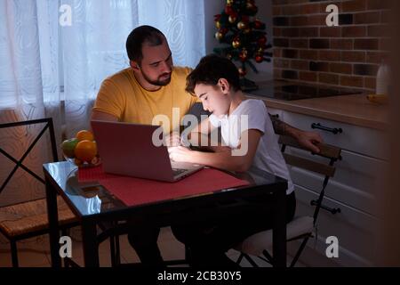 jeune caucasien attentif et prudent père aider son à étudier, à faire des mathématiques à la maison, assis dans la cuisine avec un ordinateur portable sur la table. Devoirs, école, Banque D'Images