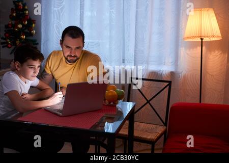 petit garçon étudiant, faisant des devoirs à la maison avant les vacances du nouvel an, un père attentif s'assoit à côté de lui et aide, en utilisant un ordinateur portable Banque D'Images