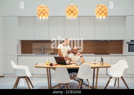 Homme concentré travaillant à la maison à l'aide d'un ordinateur portable. Asseyez-vous à la table dans la cuisine, une femme le soutient se tenir à côté de l'homme. Intérieur blanc Banque D'Images