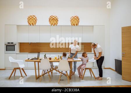 Famille caucasienne souriante dans la cuisine blanche avec intérieur moderne prenant le repas. Les enfants s'assoient à la table, les parents se tenant et les regardent. Famille, chil Banque D'Images