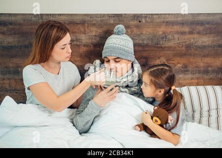 femme soigneuse et petite fille s'assoient sur le lit avec un père malade ayant une température élevée, de la fièvre. femme et enfant le soutiennent et lui donnent des médicaments Banque D'Images