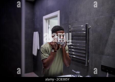 Beau homme africain mettant de la crème à raser sur son visage pendant salle de bains intérieure grise monochrome Banque D'Images