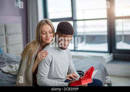 Nice dépenses de temps à la maison. Beau couple interracial de jeunes le collage de l'autre sur lit tandis que woman hugging de l'arrière un Africain ma Banque D'Images