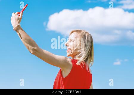 Femme blonde avec robe rouge prenant selfie avec téléphone cellulaire en bleu avec des nuages blancs ciel arrière-plan avec espace de copie pendant la journée ensoleillée d'été Banque D'Images