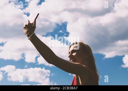 Silhouette sombre de femme blonde avec robe rouge prenant selfie avec téléphone cellulaire en bleu avec blanc nuages ciel arrière-plan avec espace de copie pendant la somme ensoleillée Banque D'Images