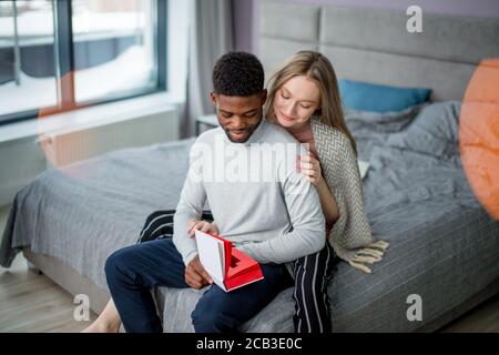 Les jeunes, de race mixte attrayant couple siéger ensemble au lit et lire un livre ensemble. Young blonde woman wearing wrap chaud sur les épaules, embrassant sa Banque D'Images