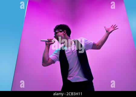 Talentueux. Jeune musicien masculin, chanteur jouant sur fond rose-bleu à la lumière du néon. Concept de musique, passe-temps, festival, divertissement, émotions. Joyeux animateur, chanteur, portrait d'artiste. Banque D'Images